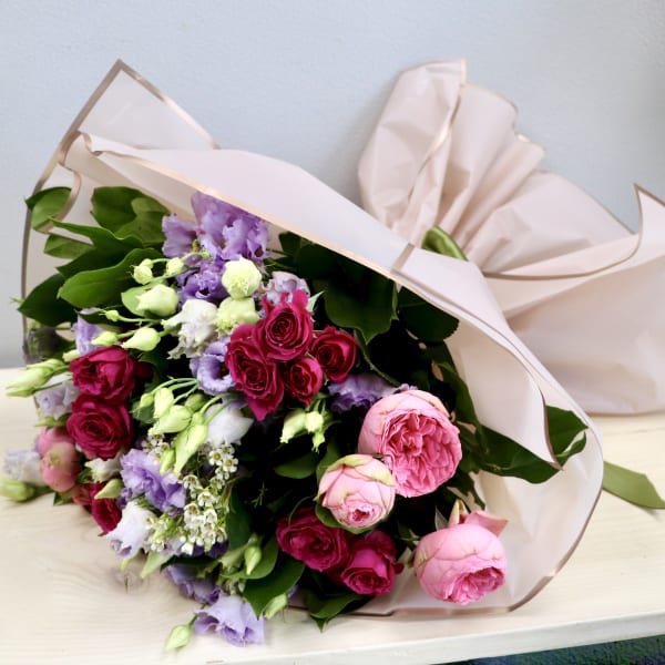 Pink and lavender bouquet of roses and lisianthus wrapped in blush paper, resting on a light wood surface