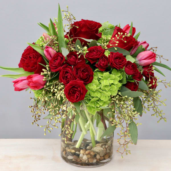 Arrangement of red roses, pink tulips, and green hydrangeas in a clear glass vase with river rocks