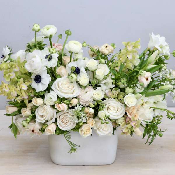 Low white and cream flower arrangement with roses, ranunculus, anemones and orchids in a rectangular white vase.