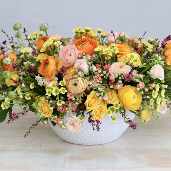 Low centerpiece of yellow, orange, and blush flowers arranged in a white ceramic bowl