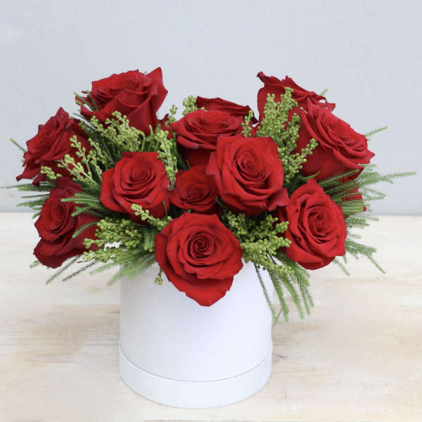 Low arrangement of red roses with green filler sprigs in a white round box.