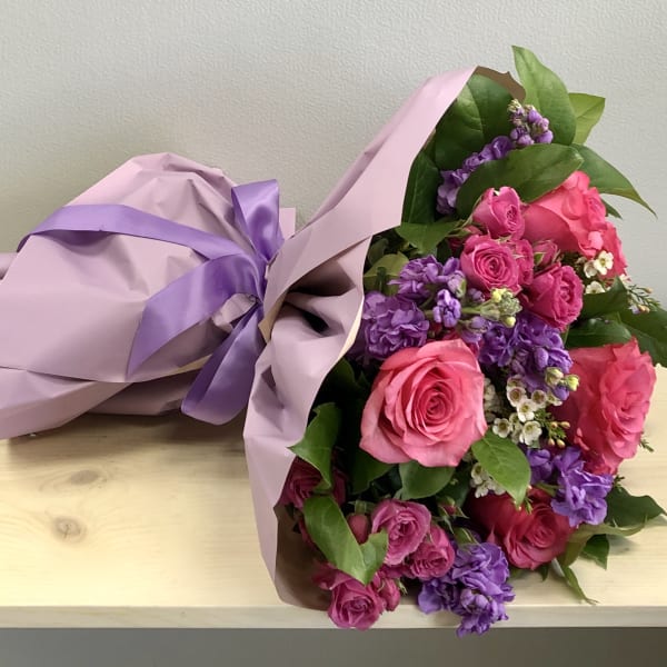 Hand-tied bouquet of pink roses and purple flowers wrapped in pink paper with a purple ribbon