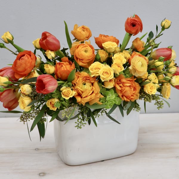 Low arrangement of orange ranunculus, yellow spray roses, and coral tulips in a white rectangular vase