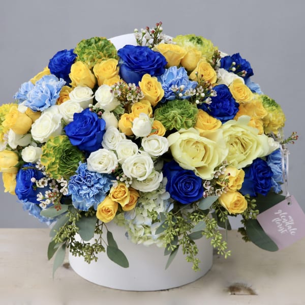 Round hatbox arrangement of yellow, blue, and white roses, carnations, and hydrangeas with a gift tag