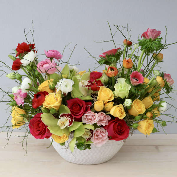 Low white bowl of mixed yellow, red, and pink roses with ranunculus and orchids in a loose, colorful arrangement
