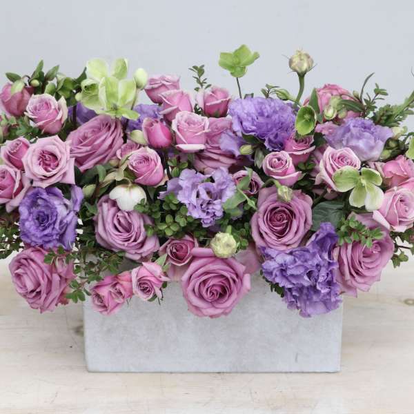 Low arrangement of pink and lavender roses with purple blooms in a rectangular white vase