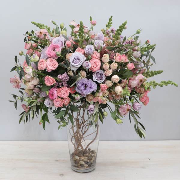 Tall arrangement of pink and lavender roses and lisianthus in a clear glass vase with stones