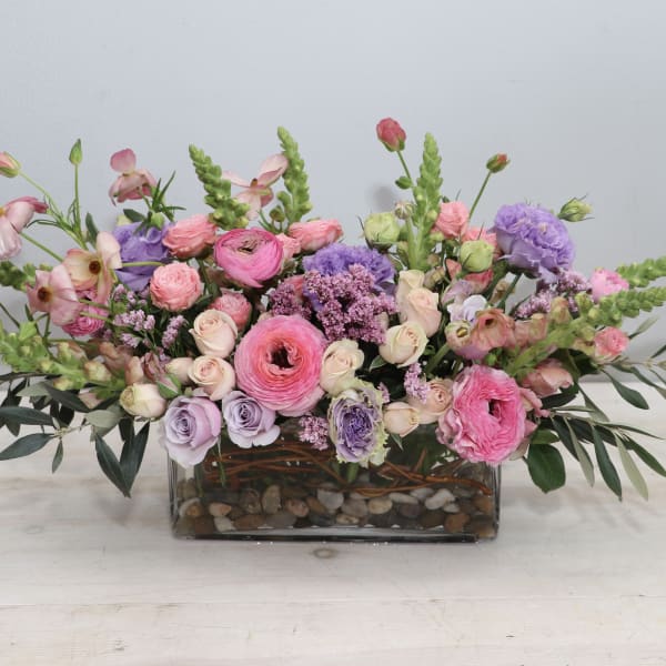 Low rectangular arrangement of pink and lavender flowers in a glass vase with river rocks