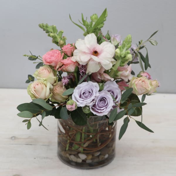 Pastel arrangement of pink and lavender roses, anemone, and snapdragons in a clear glass vase with stones