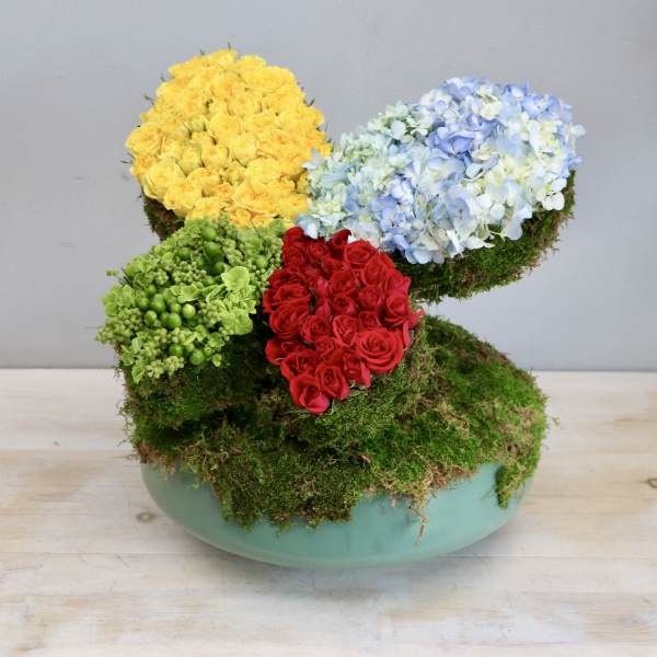Modern moss-covered arrangement with yellow and red roses and blue hydrangeas in a low green bowl