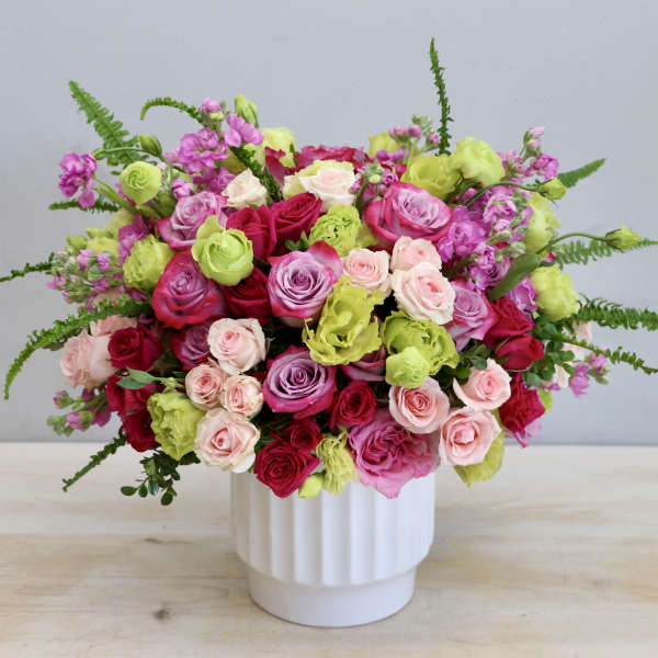 Lush arrangement of pink, red, lavender roses and green blooms in a white vase