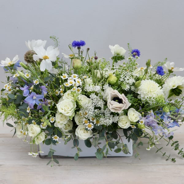 Low white ceramic centerpiece with white, blue, and purple flowers in a loose garden style