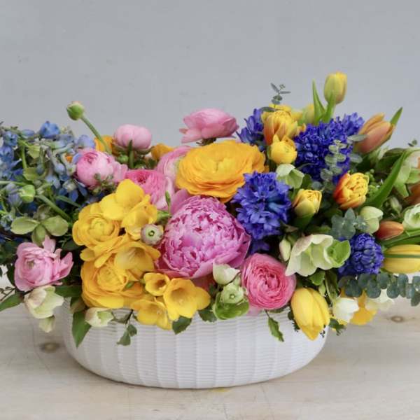 Low white bowl of bright pink, yellow, orange, and blue spring flowers in a lush arrangement