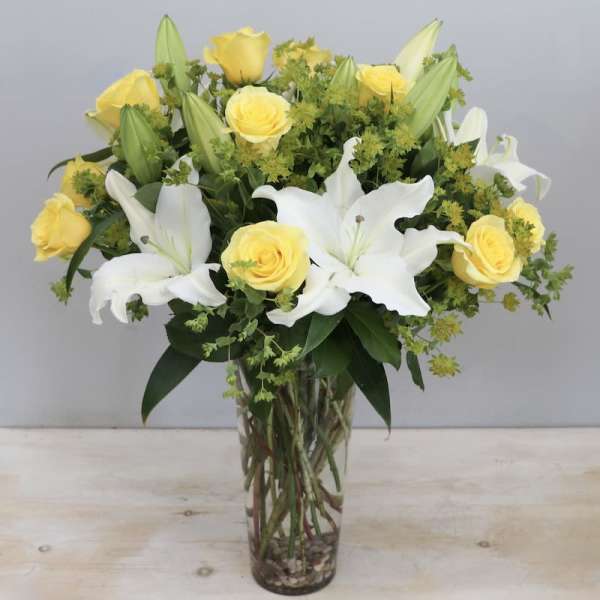 Tall glass vase of yellow roses and white lilies with green filler flowers