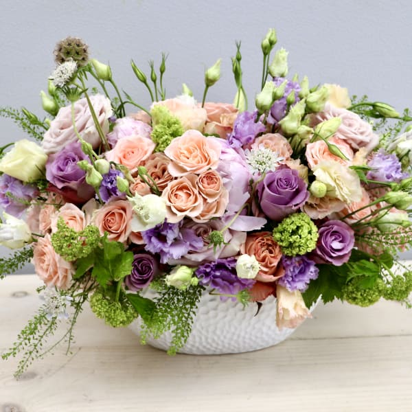 Low centerpiece with peach and lavender roses, lisianthus and green blooms in a white oval vase