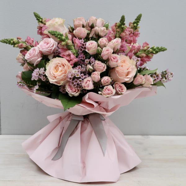 Hand-tied bouquet of light pink roses and snapdragons wrapped in pink paper with gray ribbon