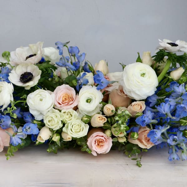 Low centerpiece of white ranunculus, blue delphinium, and blush roses in a long horizontal arrangement