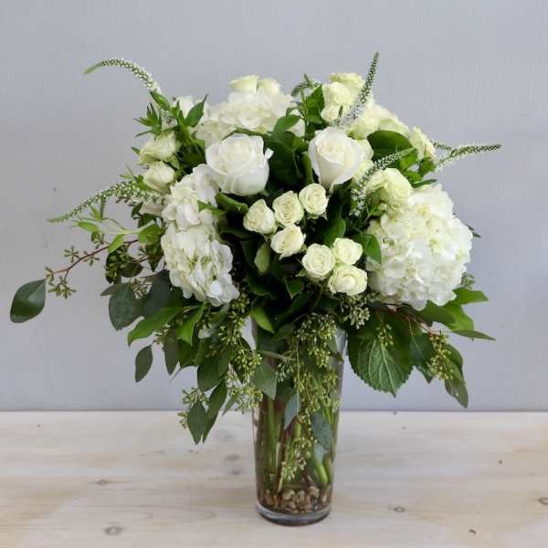 Tall arrangement of white roses and hydrangeas with trailing greenery in a clear glass vase.