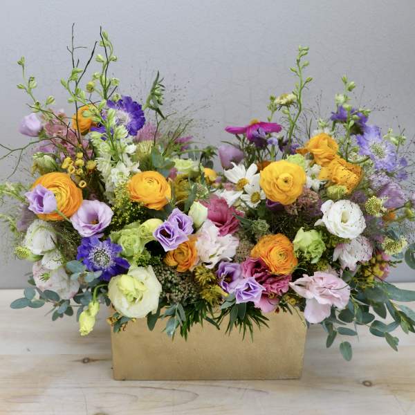 Colorful mixed flower arrangement with yellow, pink, and purple blooms in a rectangular gold container.