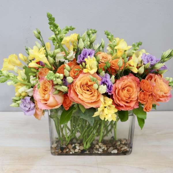 Low rectangular arrangement of orange roses, yellow flowers, and purple blooms in a glass vase with pebbles