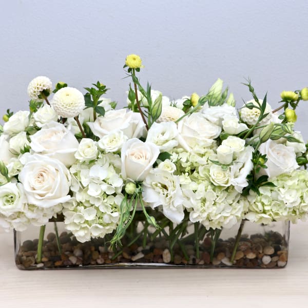 Low white rose and hydrangea arrangement in a clear rectangular vase with pebbles at the base