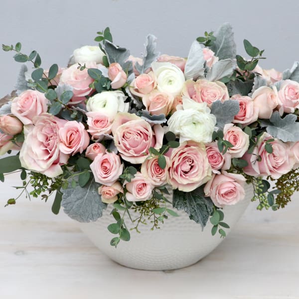 Low arrangement of blush pink roses and white blooms in a white bowl with silver foliage accents.