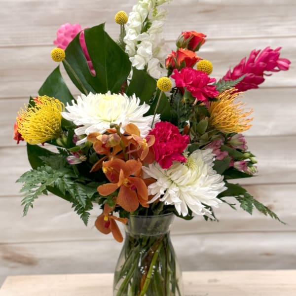 Mixed bouquet of white mums, pink carnations, yellow protea, and orange orchids in a clear glass vase
