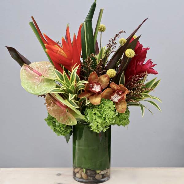 Tall tropical arrangement with red and orange blooms, orchids, and green hydrangea in a glass vase.
