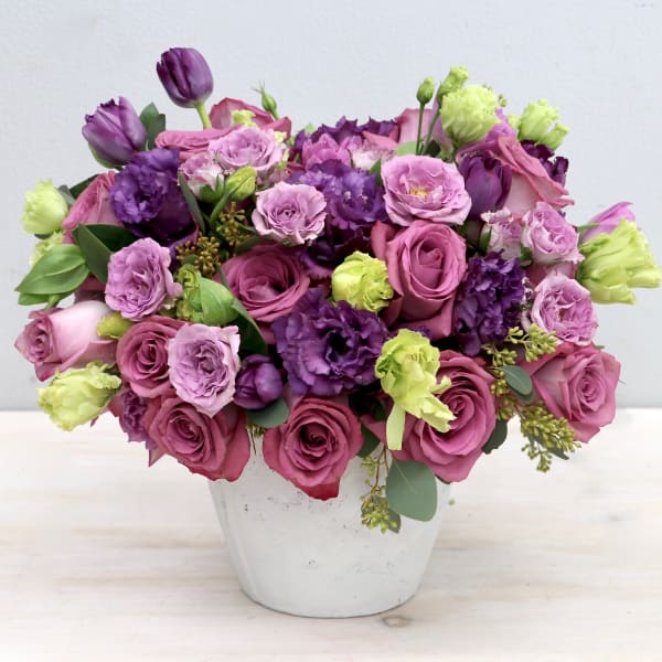 Round arrangement of mauve roses, purple lisianthus, and green blooms in a white pot