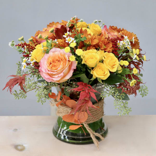 Low glass vase of yellow roses, peach rose, orange mums, and small white daisies with rustic twine wrap.