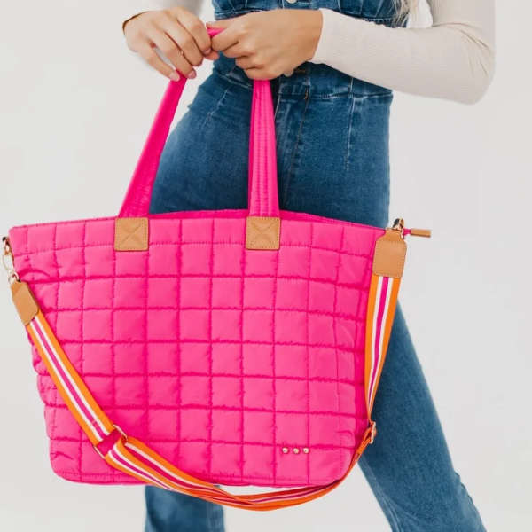 Bright pink quilted tote bag with striped shoulder strap held by a person in denim overalls.