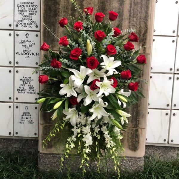 Standing spray of red roses, white lilies, and cascading orchids on an easel by a mausoleum wall