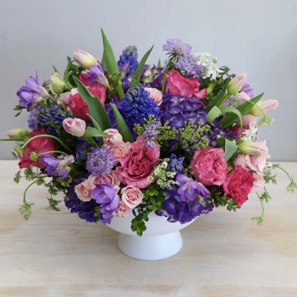 Low arrangement of pink roses, tulips, and purple blooms in a white pedestal bowl