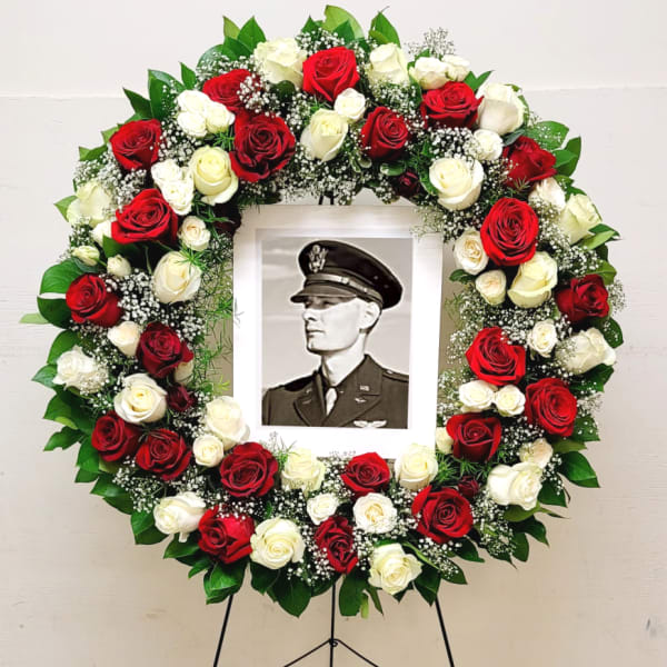 Standing circular wreath of red and white roses with baby’s breath framing a black-and-white portrait.