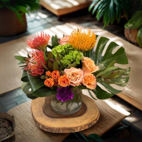 Tropical arrangement with orange protea, roses, and monstera leaves in a glass vase