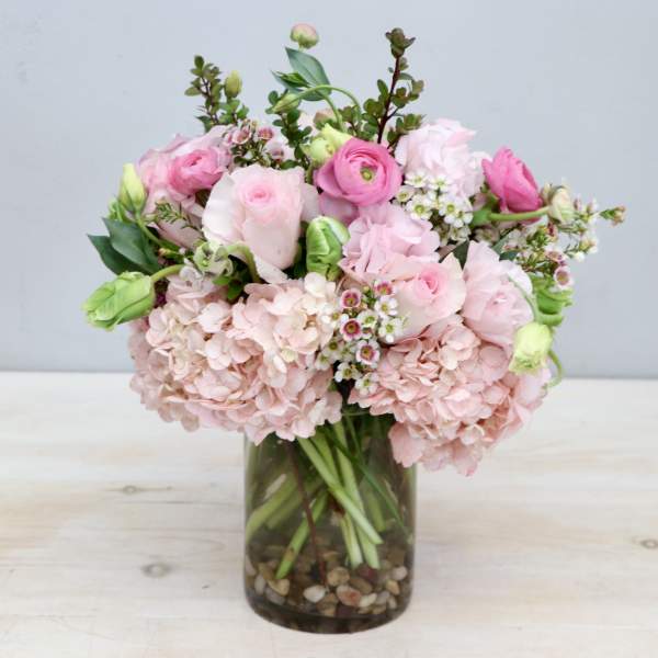 Round arrangement of pink hydrangeas, roses, and ranunculus in a clear glass vase with pebbles.