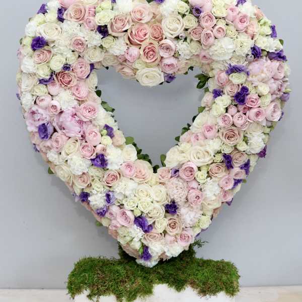 Heart-shaped standing arrangement of pink and white roses with purple accents on a moss-covered base.
