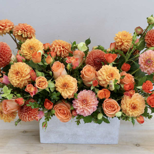 Low rectangular arrangement of orange dahlias and peach roses in a gray container