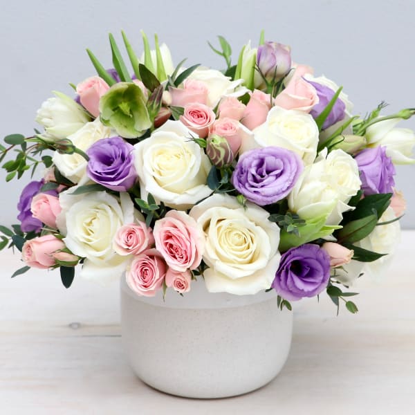 Round arrangement of white roses, pink roses, and lavender lisianthus in a white ceramic pot