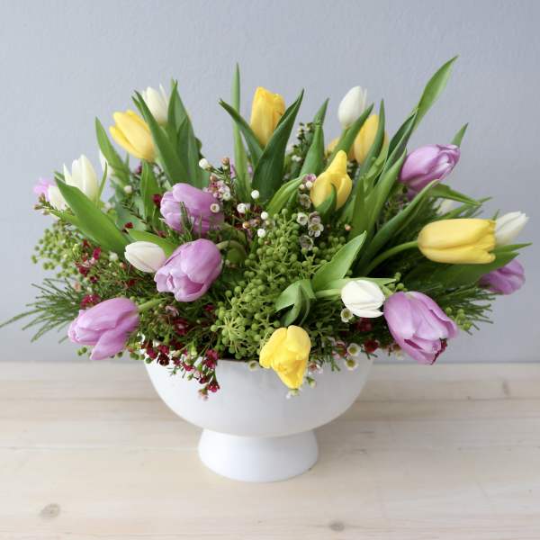 Low arrangement of yellow, white, and lavender tulips in a white pedestal bowl