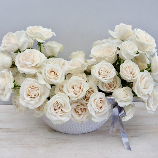 Low white bowl of clustered ivory roses with a gray ribbon accent on a light wood surface