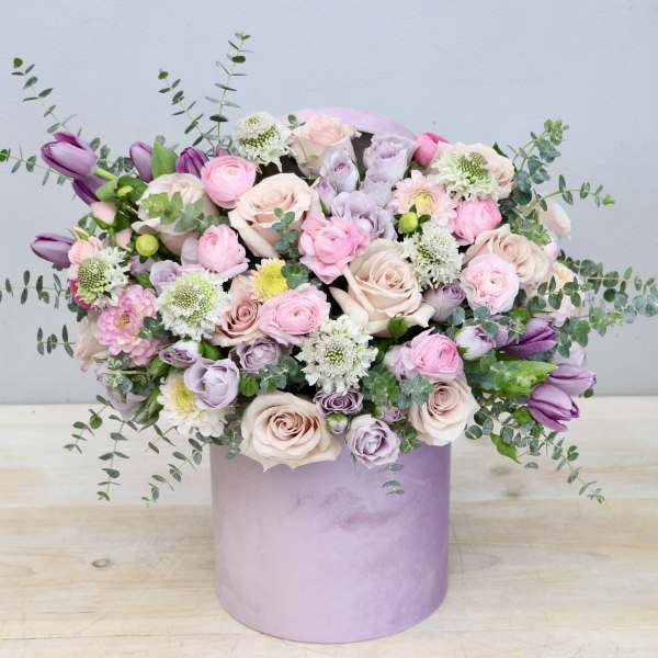 Pastel pink and lavender flower arrangement in a round lavender hatbox-style container
