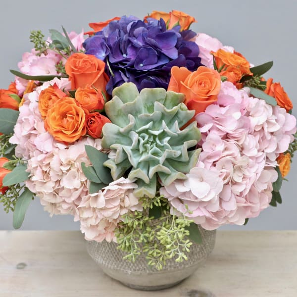 Low bowl arrangement of pink hydrangeas, orange roses, purple blooms, and a central green succulent