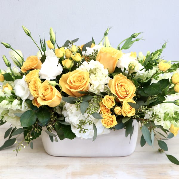 Low arrangement of yellow roses and white flowers in a rectangular white container