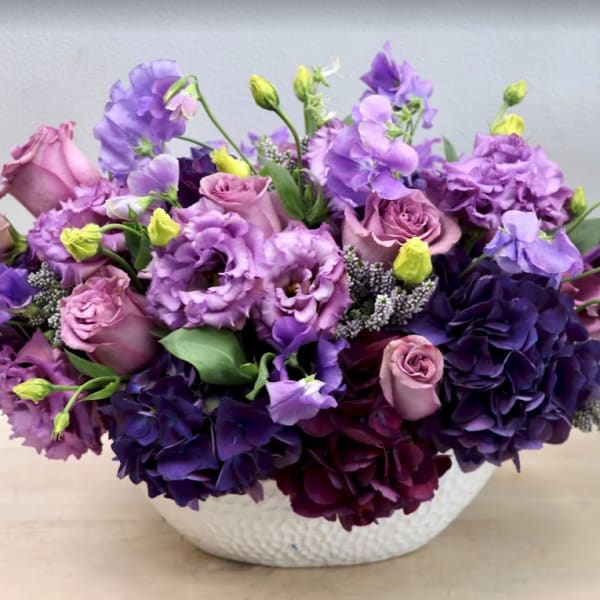 Low arrangement of purple and lavender roses and lisianthus in a white bowl vase