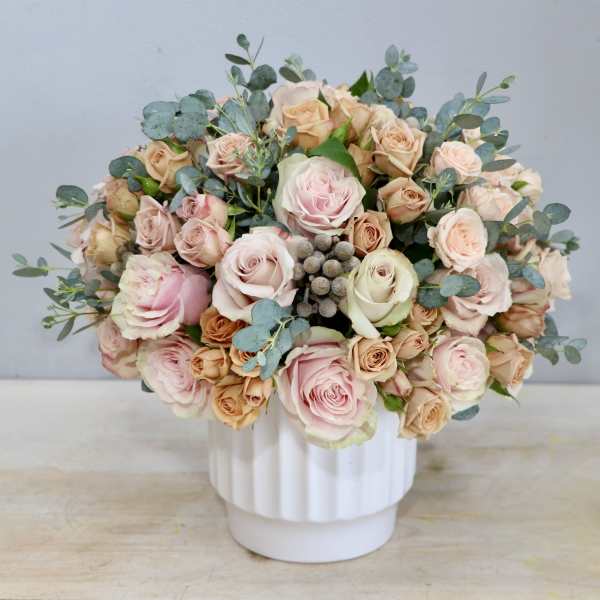 Round arrangement of blush and peach roses in a white ceramic vase with soft gray pods
