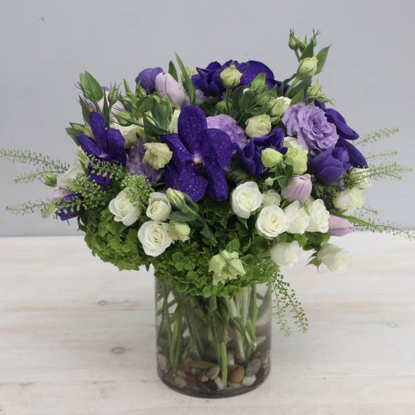 Round arrangement of purple and white flowers with green hydrangea in a clear glass cylinder vase