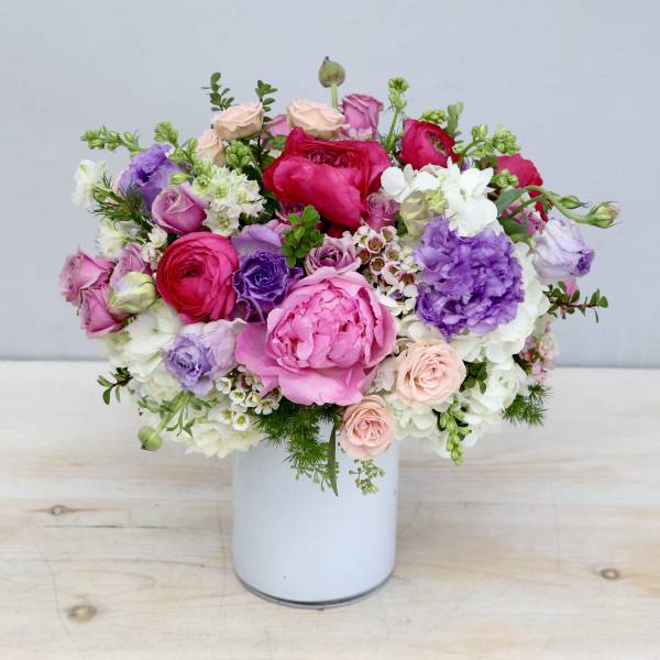 Round arrangement of pink peonies, roses, and purple blooms in a white vase