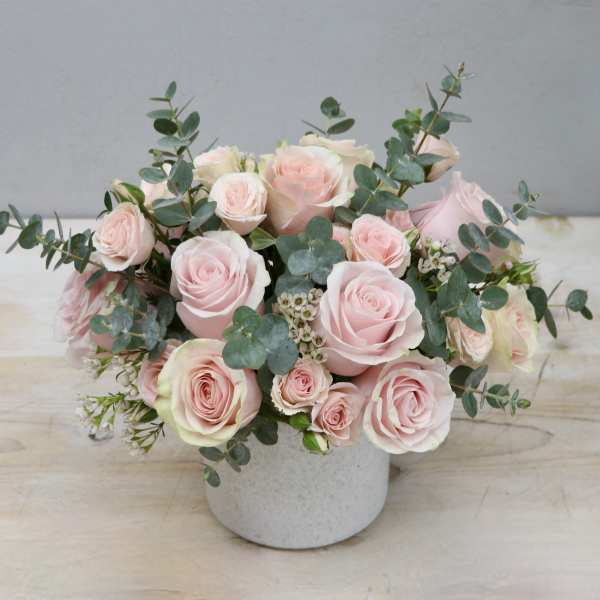 Low arrangement of pale pink roses and spray roses with eucalyptus in a white ceramic vase