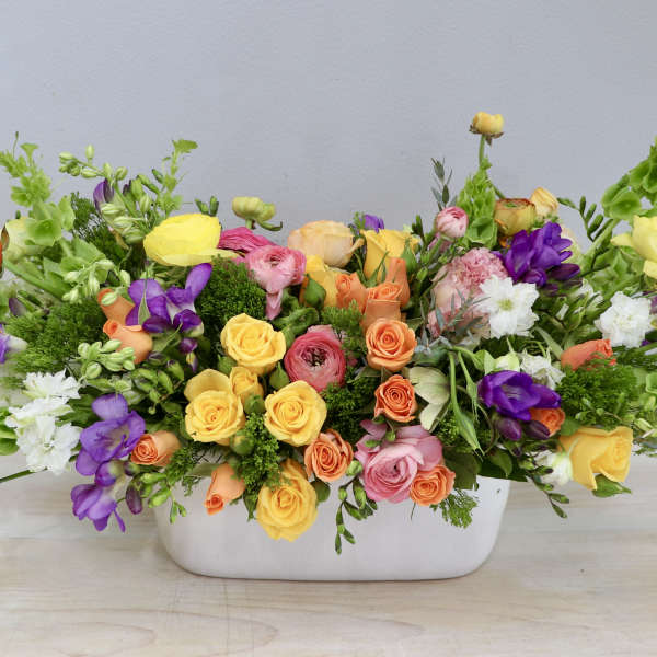 Low centerpiece of yellow, pink, orange, and purple flowers in a white rectangular container